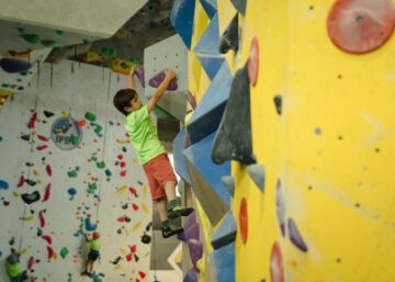 Niño escalando en muro de boulder de Climbat, con camiseta verde fluorescente, practicando en presas moradas sobre pared amarilla y azul en instalaciones indoor.