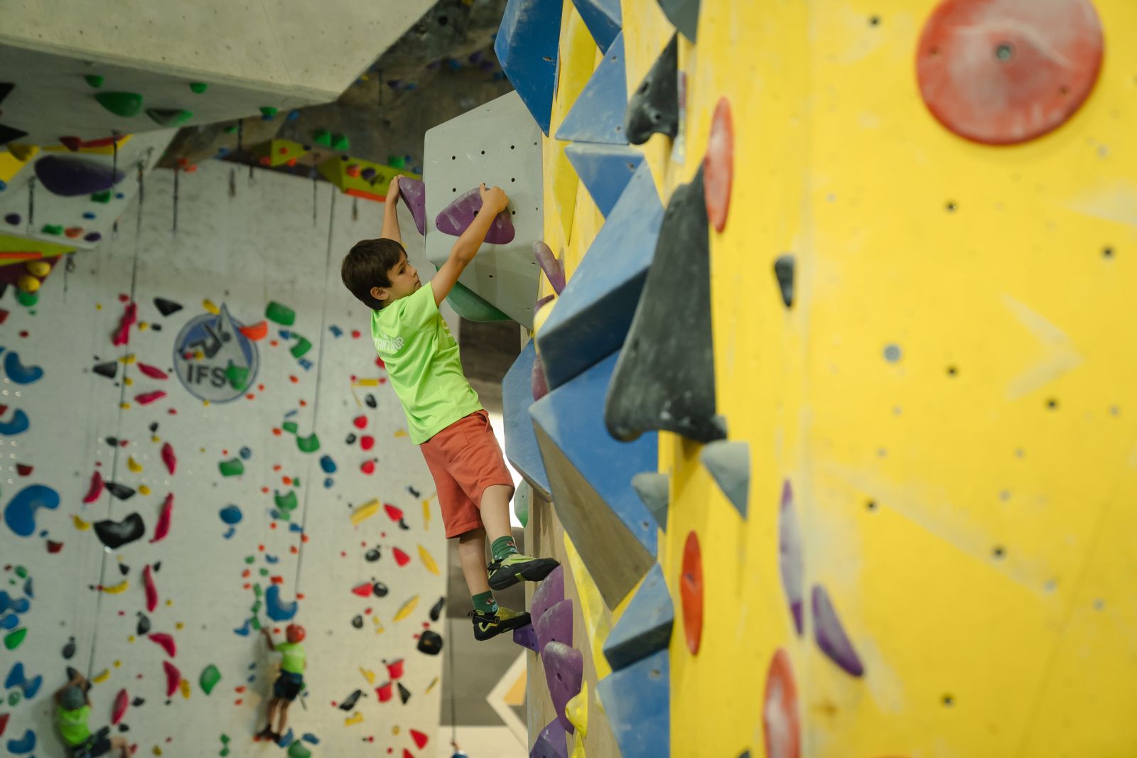 Niño escalando en muro de boulder de Climbat, con camiseta verde fluorescente, practicando en presas moradas sobre pared amarilla y azul en instalaciones indoor.