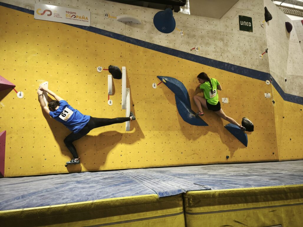 Dos escaladores universitarios compitiendo simultáneamente en la modalidad de bloque durante el Campeonato de España Universitario de Escalada 2025 en Climbat X-Madrid. El competidor con dorsal 61 en camiseta azul realiza un movimiento de apertura, mientras la escaladora con dorsal 30 en camiseta verde fluorescente asciende por un volumen azul.