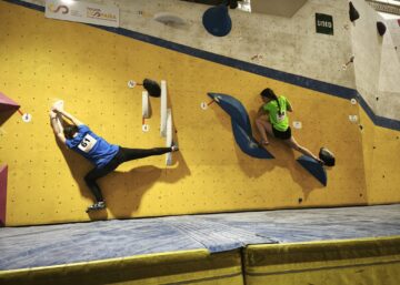 Dos escaladores universitarios compitiendo simultáneamente en la modalidad de bloque durante el Campeonato de España Universitario de Escalada 2025 en Climbat X-Madrid. El competidor con dorsal 61 en camiseta azul realiza un movimiento de apertura, mientras la escaladora con dorsal 30 en camiseta verde fluorescente asciende por un volumen azul.